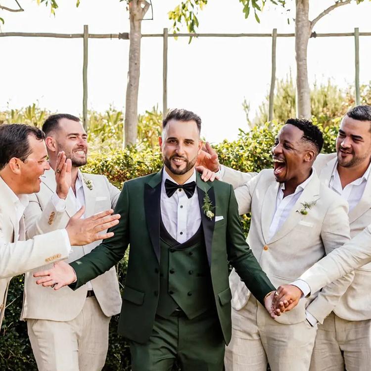 A groom in a deep green three piece suit surrounded by his groomsmen in linen